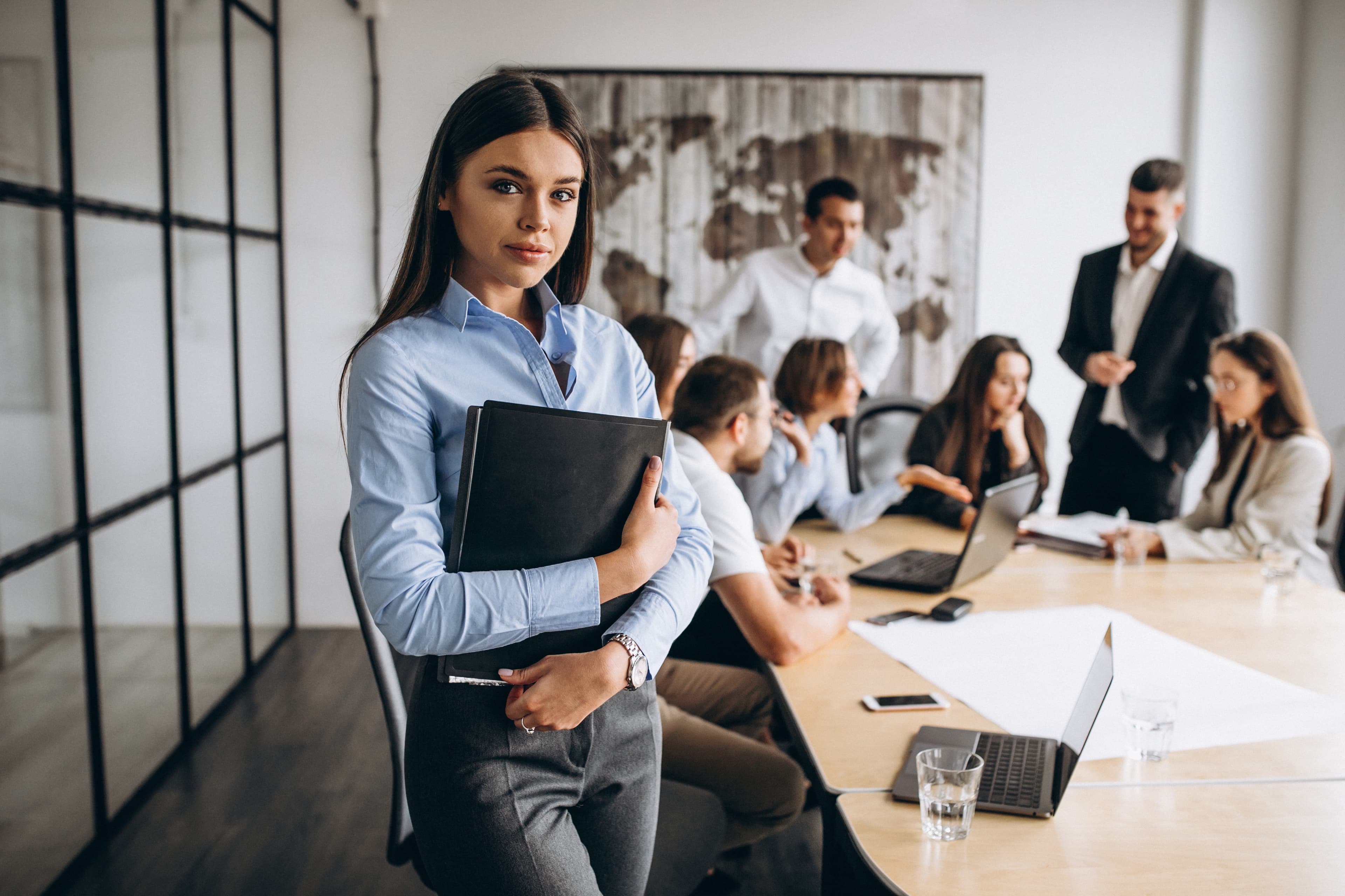 Professional and confident looking woman facing the user while in the back other coworkers people are working colaboratively
